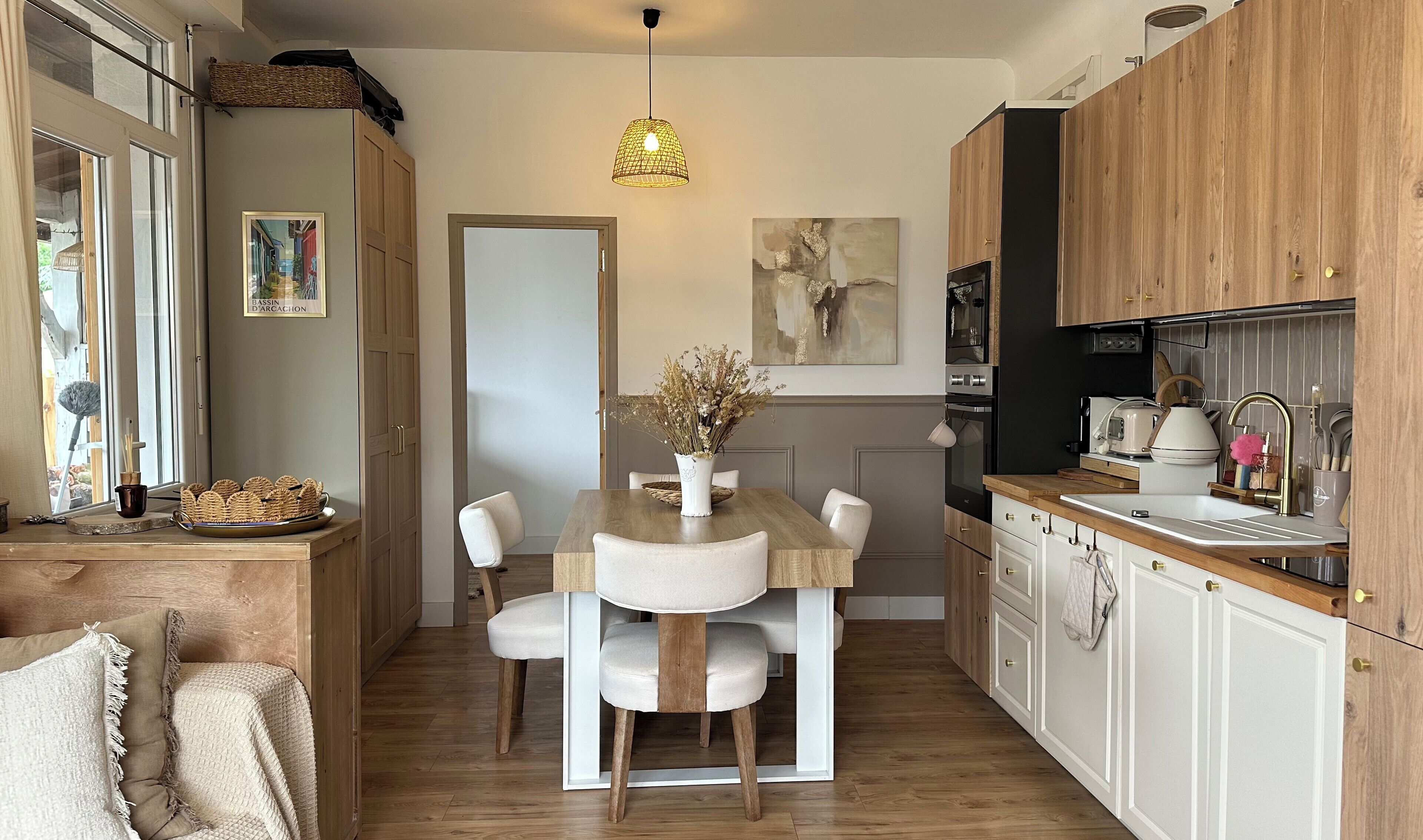 Een moderne keuken met houten kastjes, een eettafel met stoelen, een hanglamp en een schilderij aan de muur.