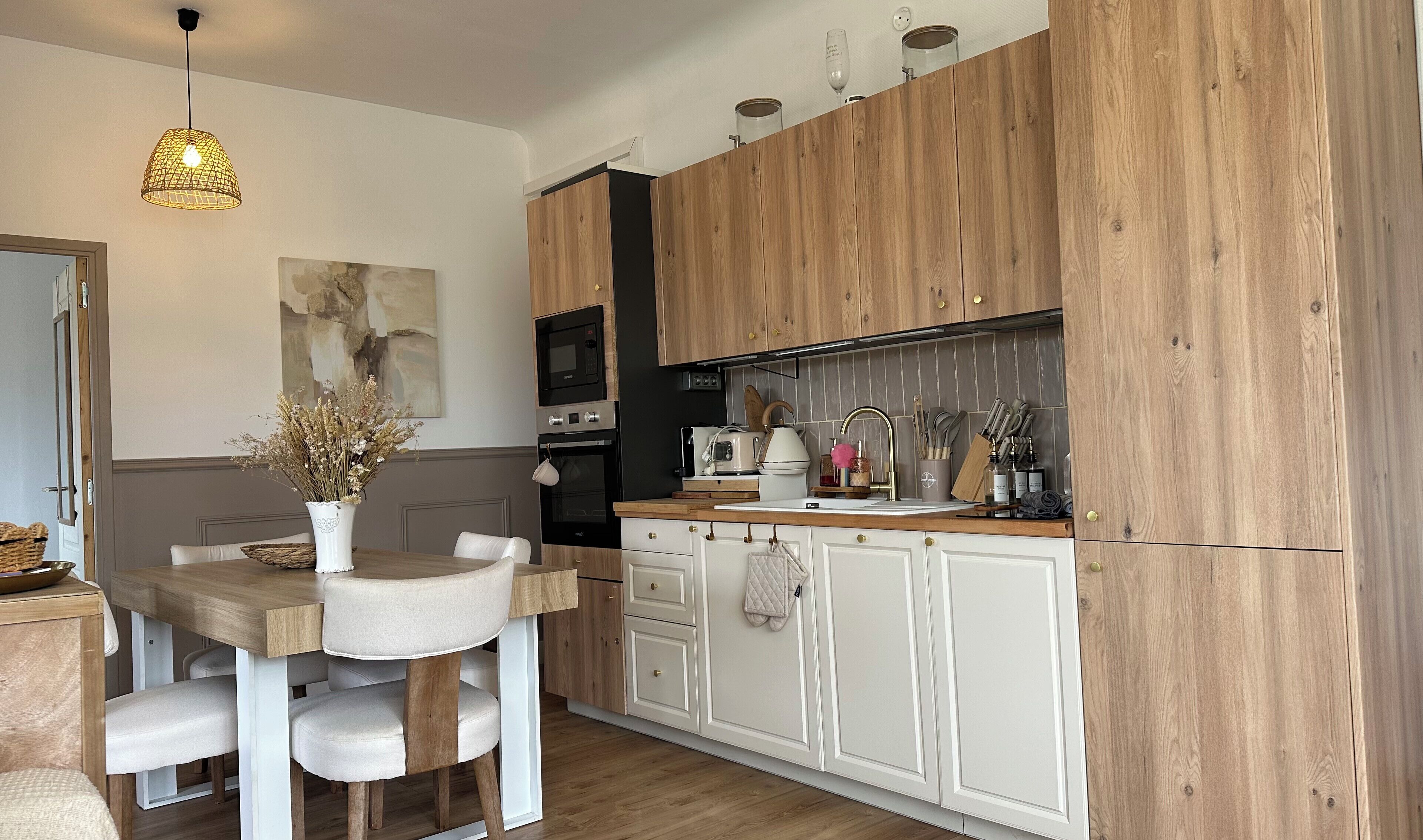Een moderne keuken met houten kastjes, een eettafel met stoelen, een hanglamp en een vaas met gedroogde bloemen.