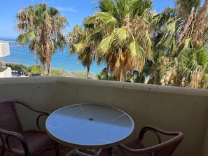 A balcony with a round table and two wicker chairs, overlooking a coastal area with palm trees and a view of the sea. - Apartment 'Chris' with Shared Pool, Wi-Fi, and Air Conditioning (Benalmádena)