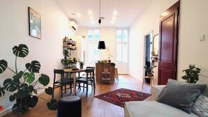 Een gezellige woonkamer met een bank, een eettafel met stoelen, een bijzettafeltje en een klein vloerkleed. Rechts is een deur en links een raam.
