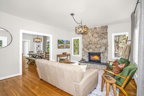 Ein Wohnzimmer mit Kamin aus Stein, einer Couch, einem Sessel und einem Essbereich mit Tisch und Stühlen.