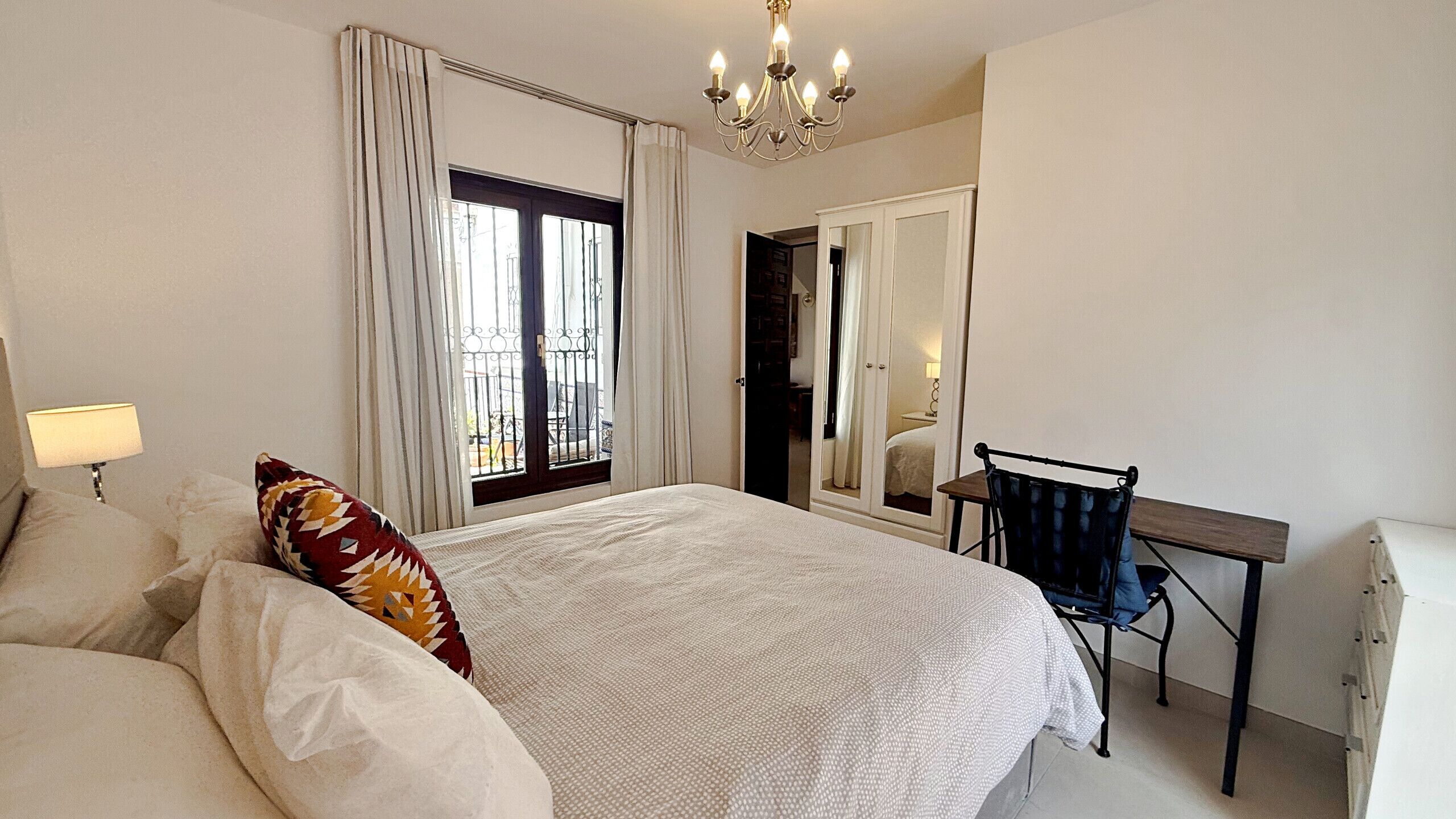 A bedroom with a bed, a desk, a chair, a chandelier, and a window with curtains.