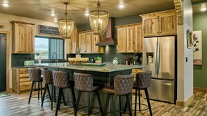 A kitchen with wooden cabinets, a central island with bar stools, a stainless steel refrigerator, and pendant lighting. - RiverBend Rio (South Fork)
