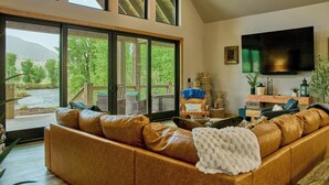 A spacious living room with a large leather sofa, a flat-screen TV, and a view of a deck and trees. - RiverBend Rio (South Fork)