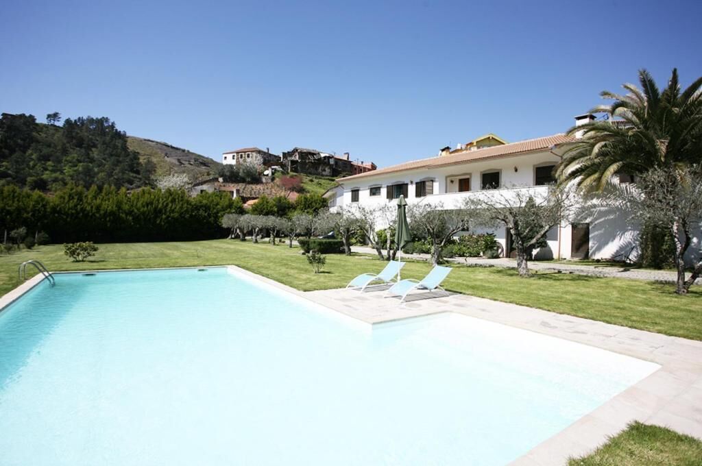 Una piscina amb gandules, envoltada per un jardí ben cuidat i un edifici amb teulada de rajola.