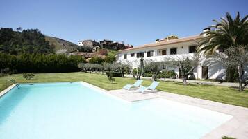 Una piscina amb gandules, envoltada per un jardí ben cuidat i un edifici amb teulada de rajola.