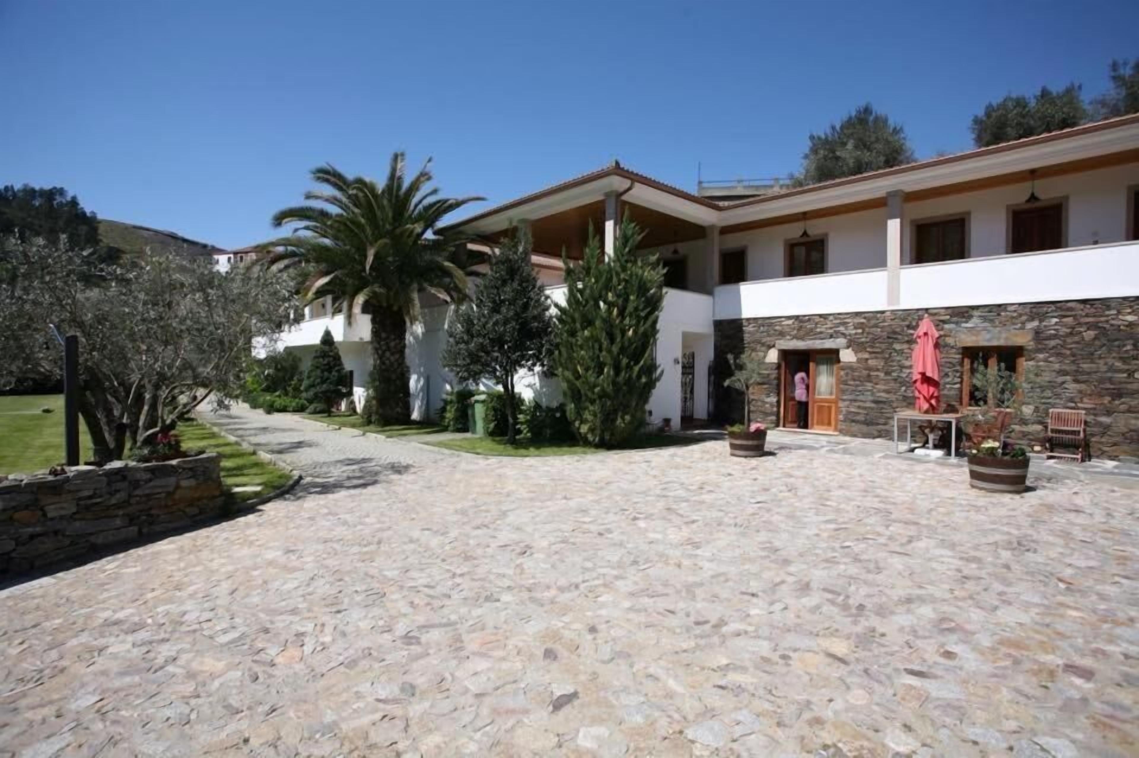 A stone-paved courtyard with a building featuring a stone facade and wooden beams, surrounded by greenery and a clear sky.