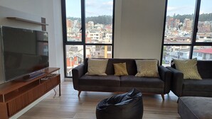 Een woonkamer met een grote televisie, twee banken, een relaxfauteuil en uitzicht op een stadsbeeld door grote ramen.
