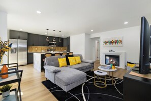 A modern living room with a grey sofa, a small coffee table, and a TV. There is a kitchen area with dark cabinets and a microwave. - Stylish Eastside 3BR Family Home - Prime Location! (Bellevue)