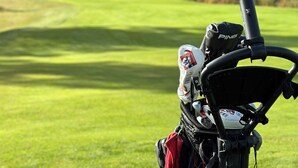 Un carrito de golf con una toalla roja y un casco en el asiento, estacionado en un campo de golf con una colina boscosa al fondo.