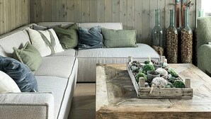 Un salón acogedor con piso de madera, un sofá gris con cojines, una mesa de centro con objetos decorativos y una lámpara grande.