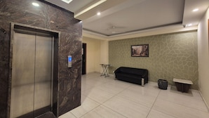 Een ontvangsthal met een marmeren lift, een zwarte bank, een kleine tafel en een ingelijste foto aan de muur.