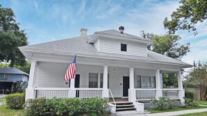 Een wit huis met twee verdiepingen, een veranda, een Amerikaanse vlag en een schoorsteen.
