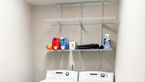 Une buanderie équipée de deux machines à laver et de deux sèche-linge à chargement frontal, tous deux de couleur blanche. Au-dessus, une étagère en métal contient les produits de nettoyage, et un panneau en bois orne le mur gauche.