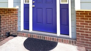 Une porte bleue avec des moulures blanches, un paillasson noir et une lanterne suspendue.