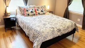 Une chambre à coucher avec un lit, deux tables de chevet, une commode, une lampe et une fenêtre avec des rideaux.