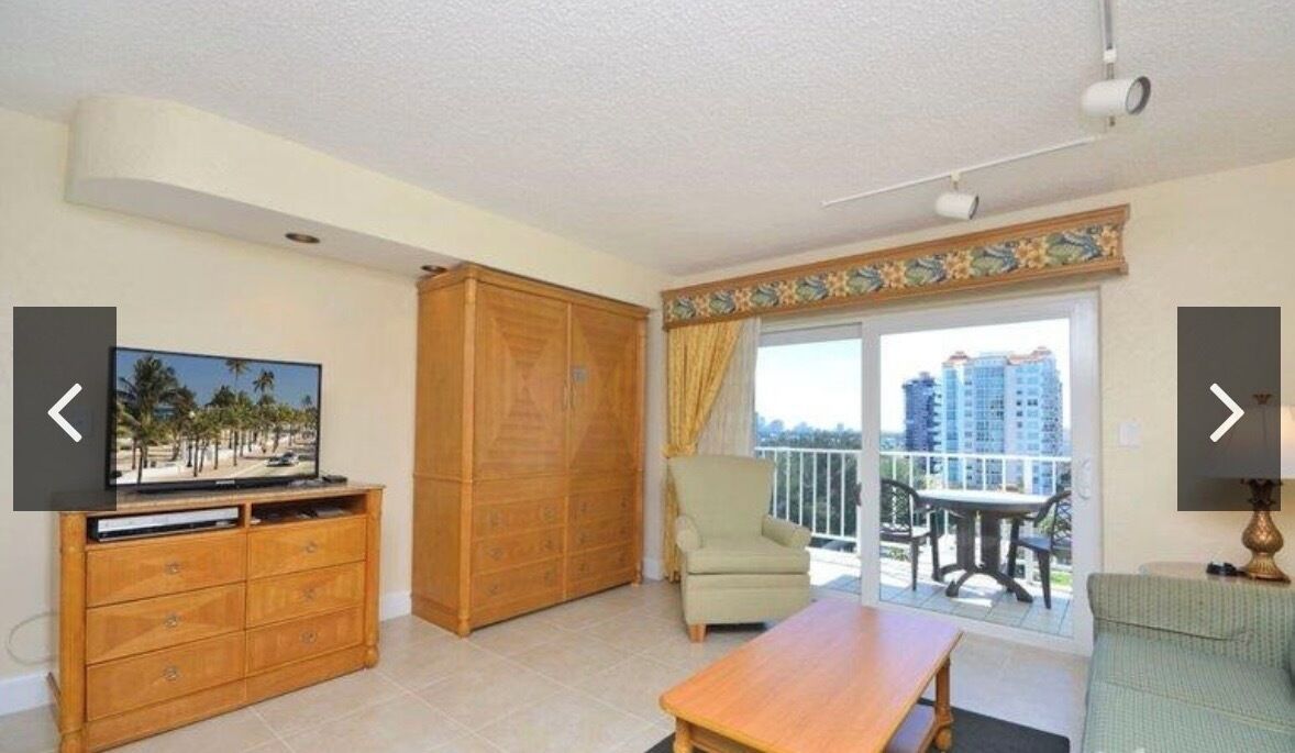 A room with a TV, wooden cabinet, armchair, table, and balcony with a view of buildings.