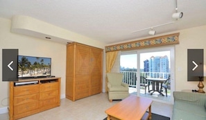 Ein Zimmer mit Fernseher, Holzschrank, Sessel, Tisch und einem Balkon mit Blick auf Gebäude.