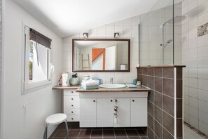 Ein Badezimmer mit weißem Waschtisch, einem Spiegel, einer Duschkabine und einem Fenster mit Jalousien.