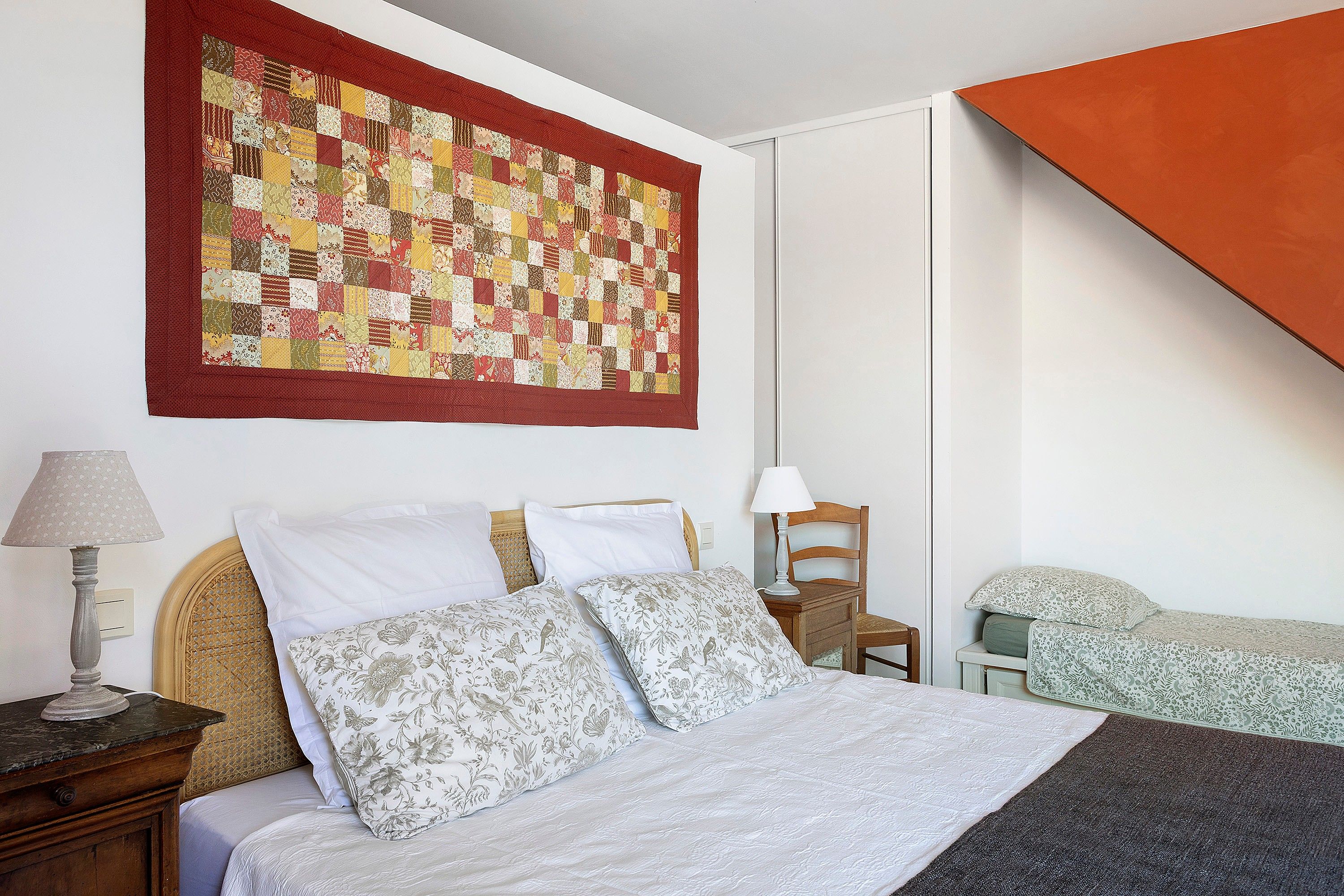 A bedroom with a bed, pillows, a lamp, a chair, and a large quilt hanging on the wall.