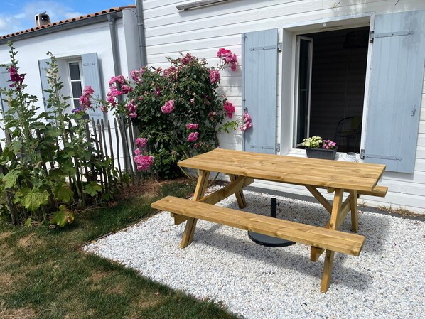 A wooden picnic table with benches in front of a white building with blue shutters.