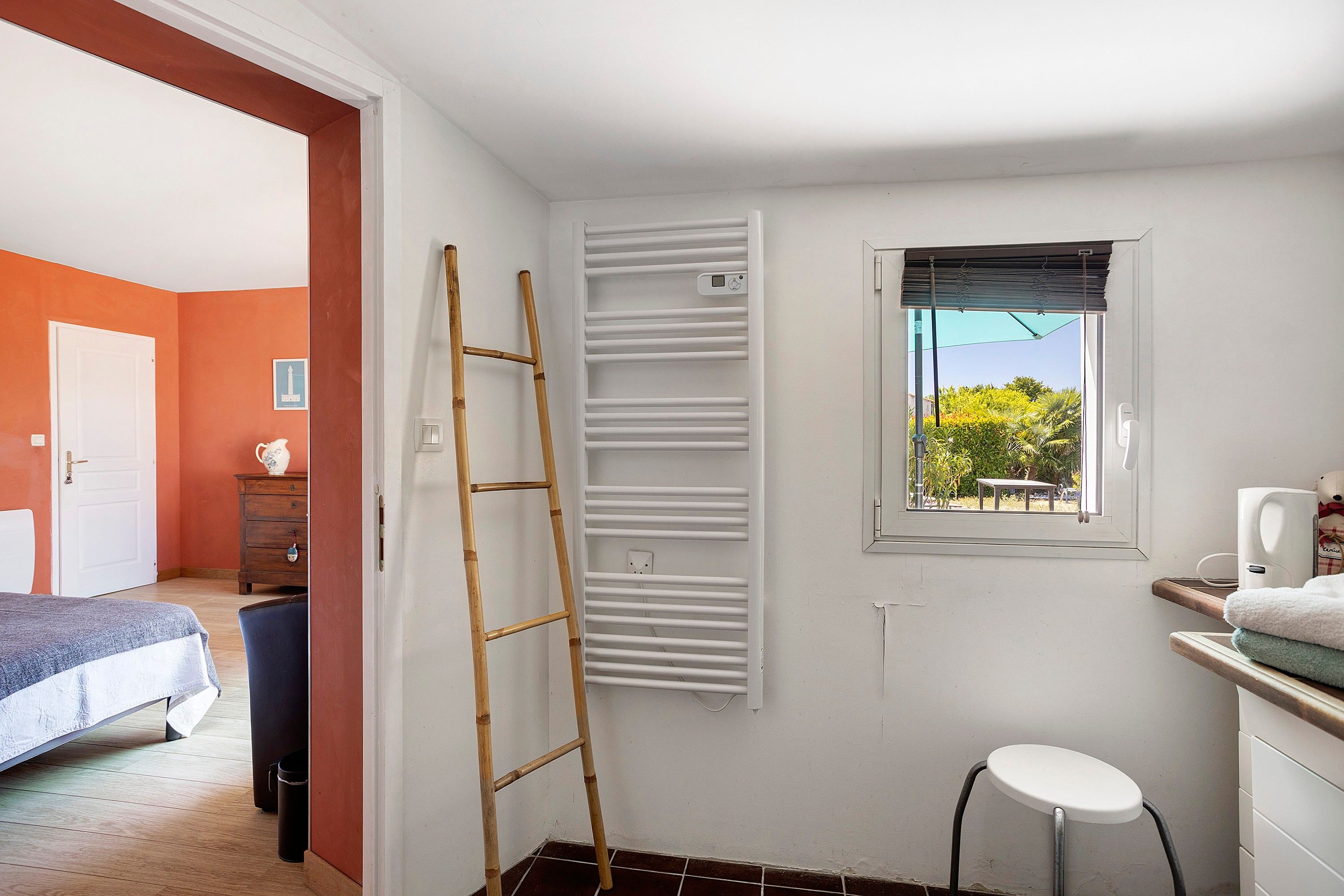 Una habitación con cama, escritorio y silla, ventana con persianas, radiador y una escalera apoyada en la pared.