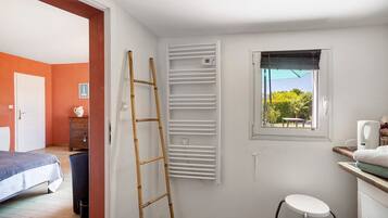 Una habitación con cama, escritorio y silla, ventana con persianas, radiador y una escalera apoyada en la pared.