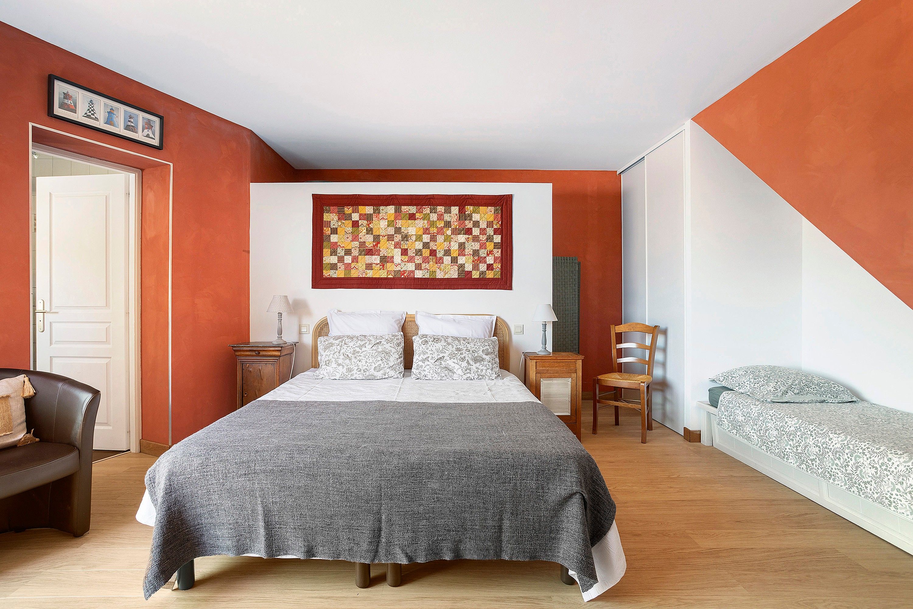 A bedroom with a large bed, a chair, a bedside table, and a wall-mounted quilt.