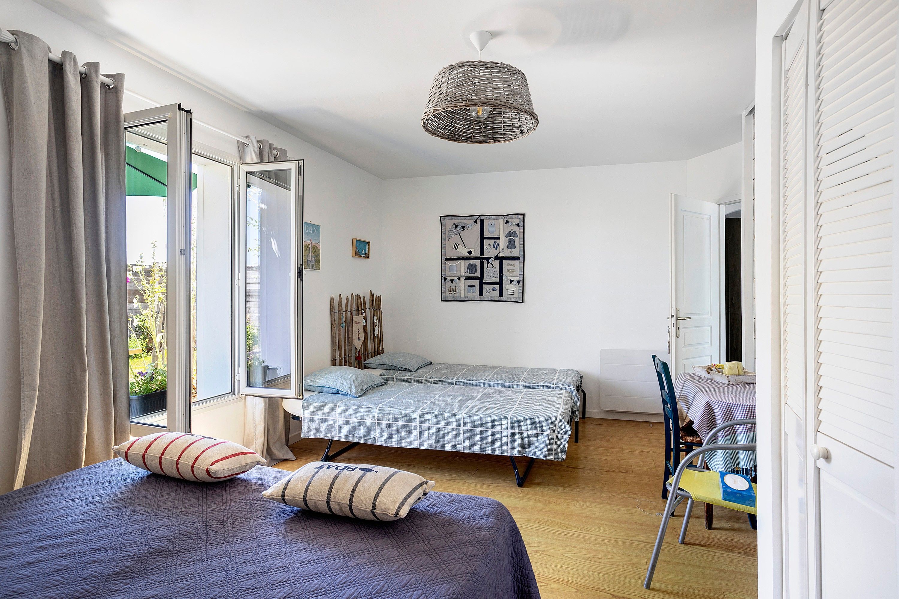 A bedroom with two beds, a window with curtains, a hanging light fixture, and a wall with a quilted artwork.