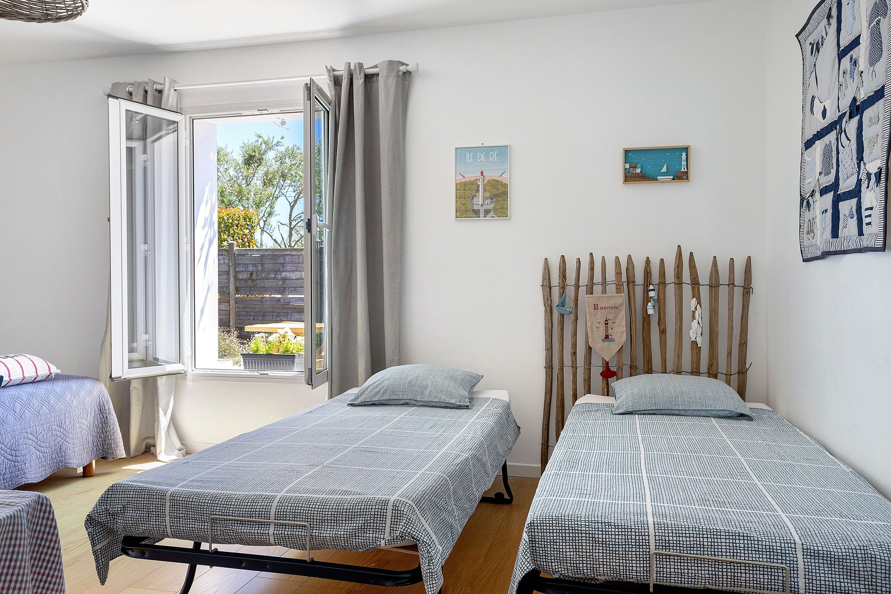 A room with two single beds, a window with curtains, a wooden fence outside, and a wall with a framed picture.