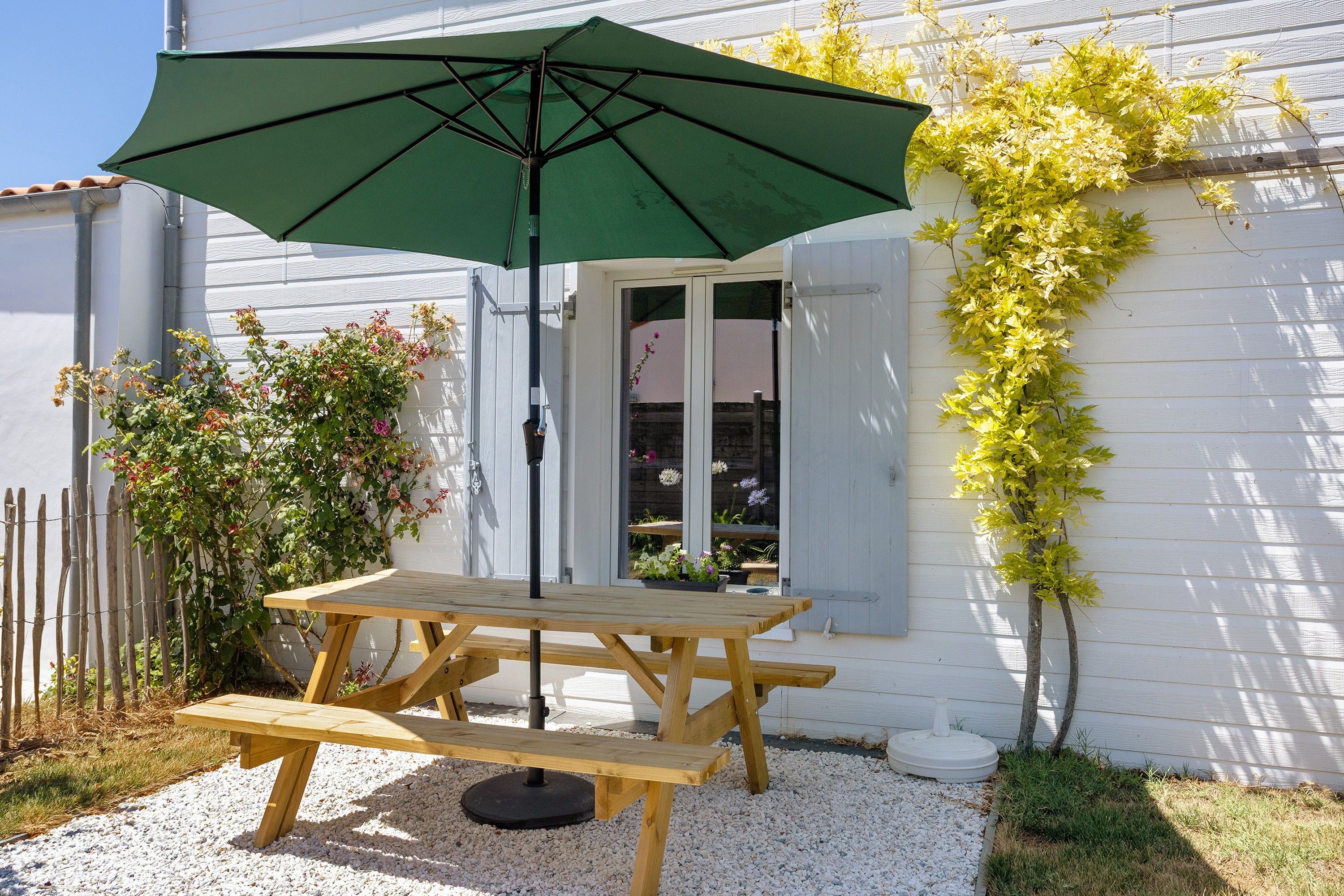 A wooden picnic table with a green umbrella, a white building with a window, and a climbing plant on the wall.