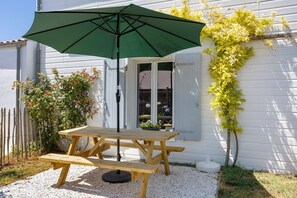 A wooden picnic table with a green umbrella, a white building with a window, and a climbing plant on the wall.