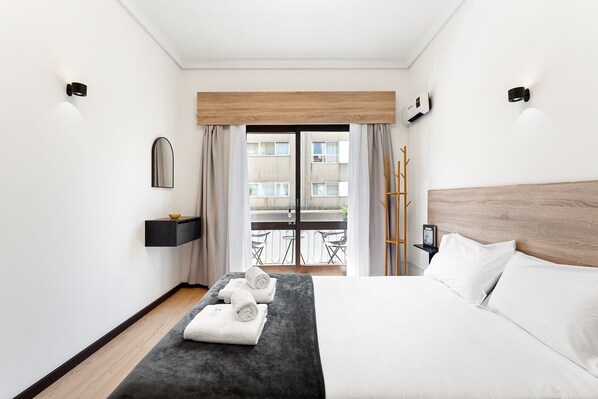 A modern hotel room with a bed, a small bedside table, a mirror, a view of the outside balcony, and a wooden headboard. - GuestReady - Oporto Breiner Terrace (Porto)