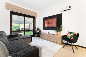 A modern living room with a large window, a flat-screen TV, a sofa, a chair, and a wooden cabinet. - GuestReady - Oporto Breiner Terrace (Porto)
