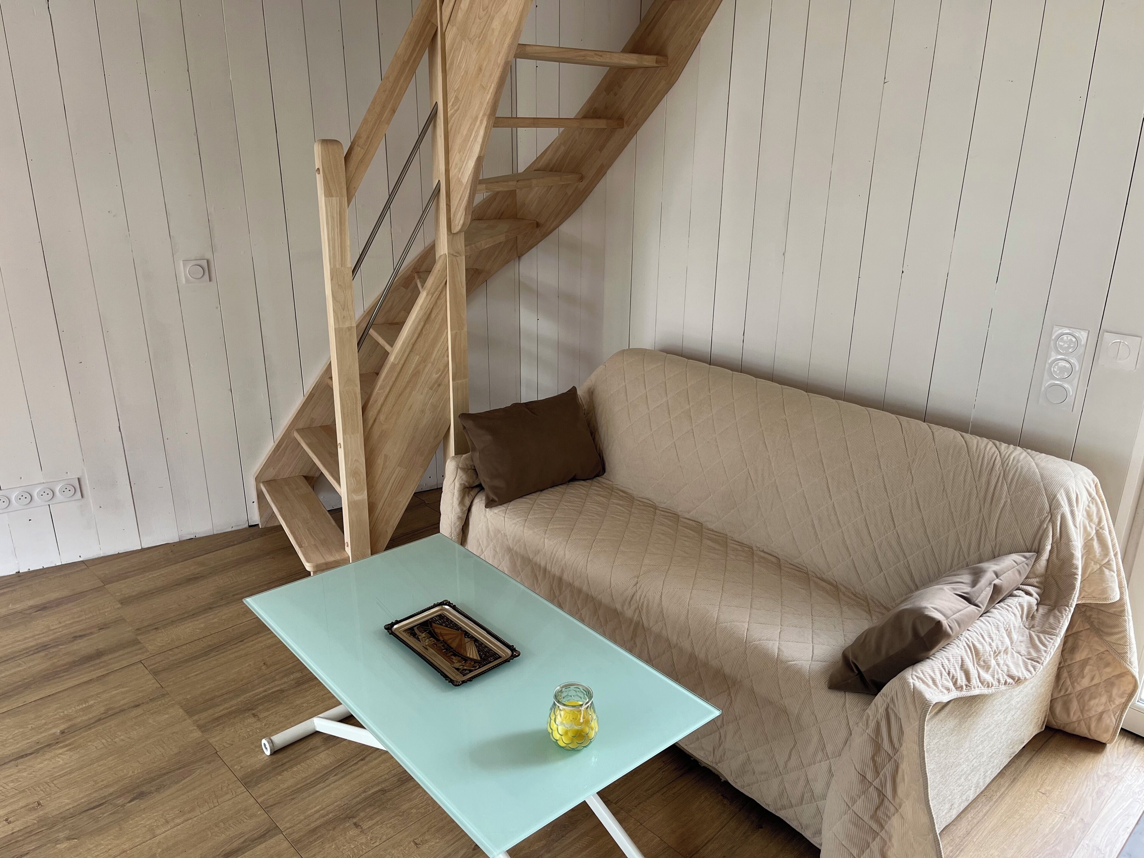A room with a wooden staircase, a beige sofa, a glass table with a picture frame and a glass jar, and wooden flooring.