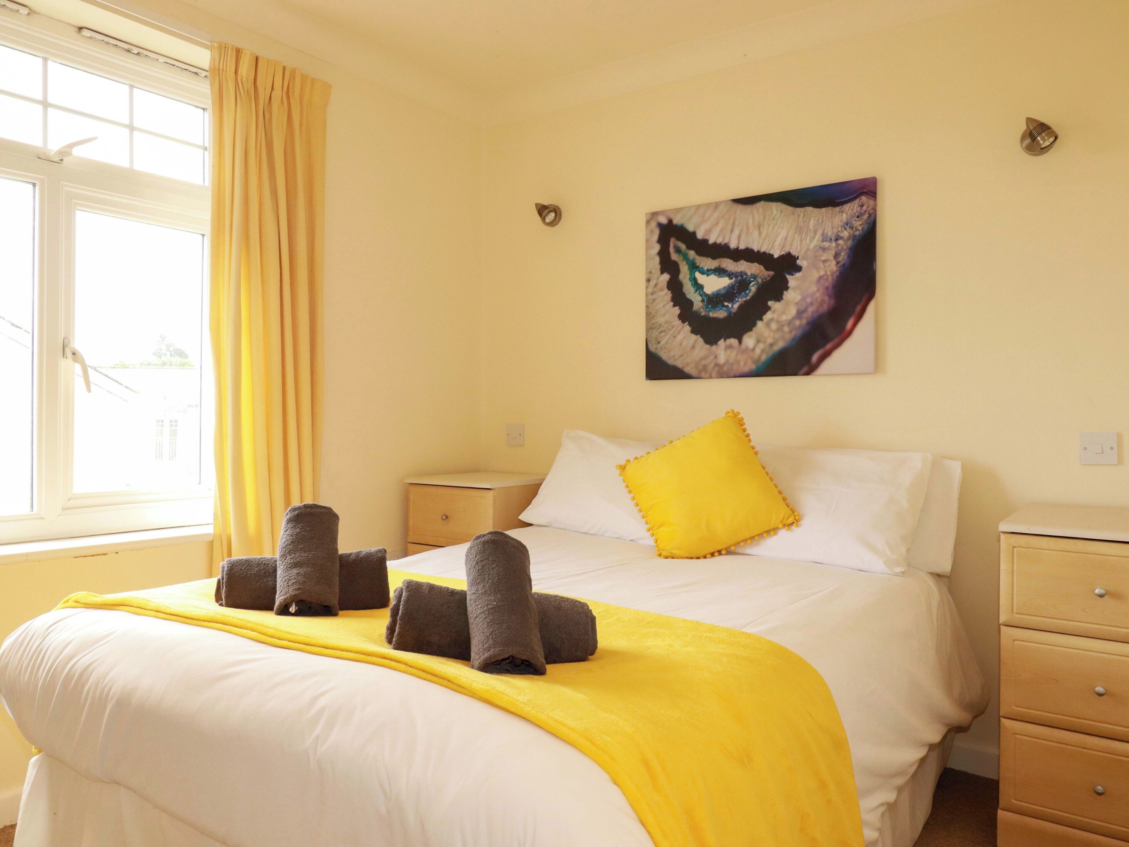 A hotel room with a bed, a nightstand, a drawer unit, a window with curtains, and a wall-mounted artwork.