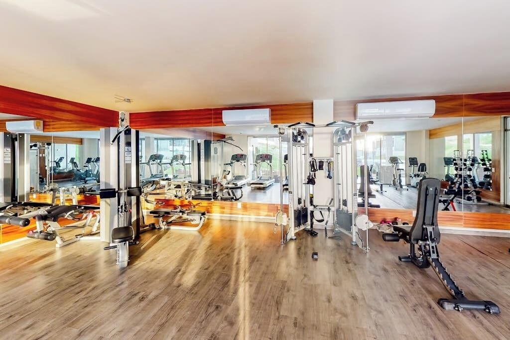 Un gimnasio con diversas máquinas de ejercicio, suelo de madera y grandes ventanales que dejan entrar luz natural.