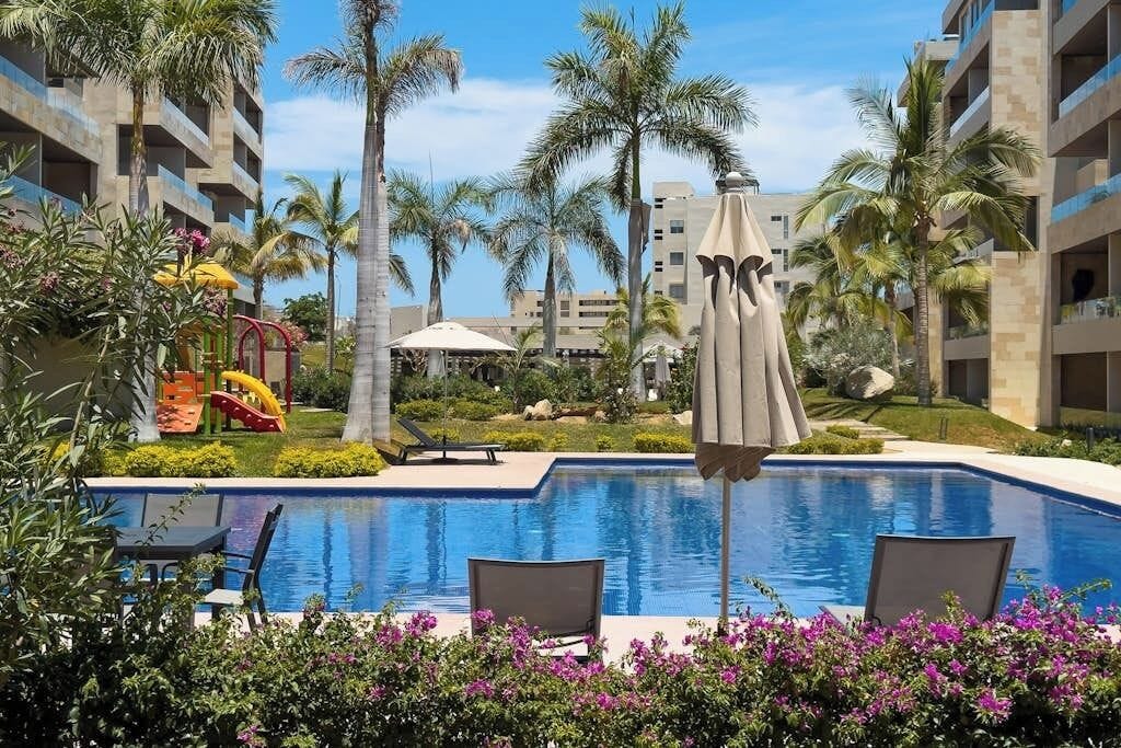 Piscina com espreguiçadeiras, palmeiras e um escorregador.
