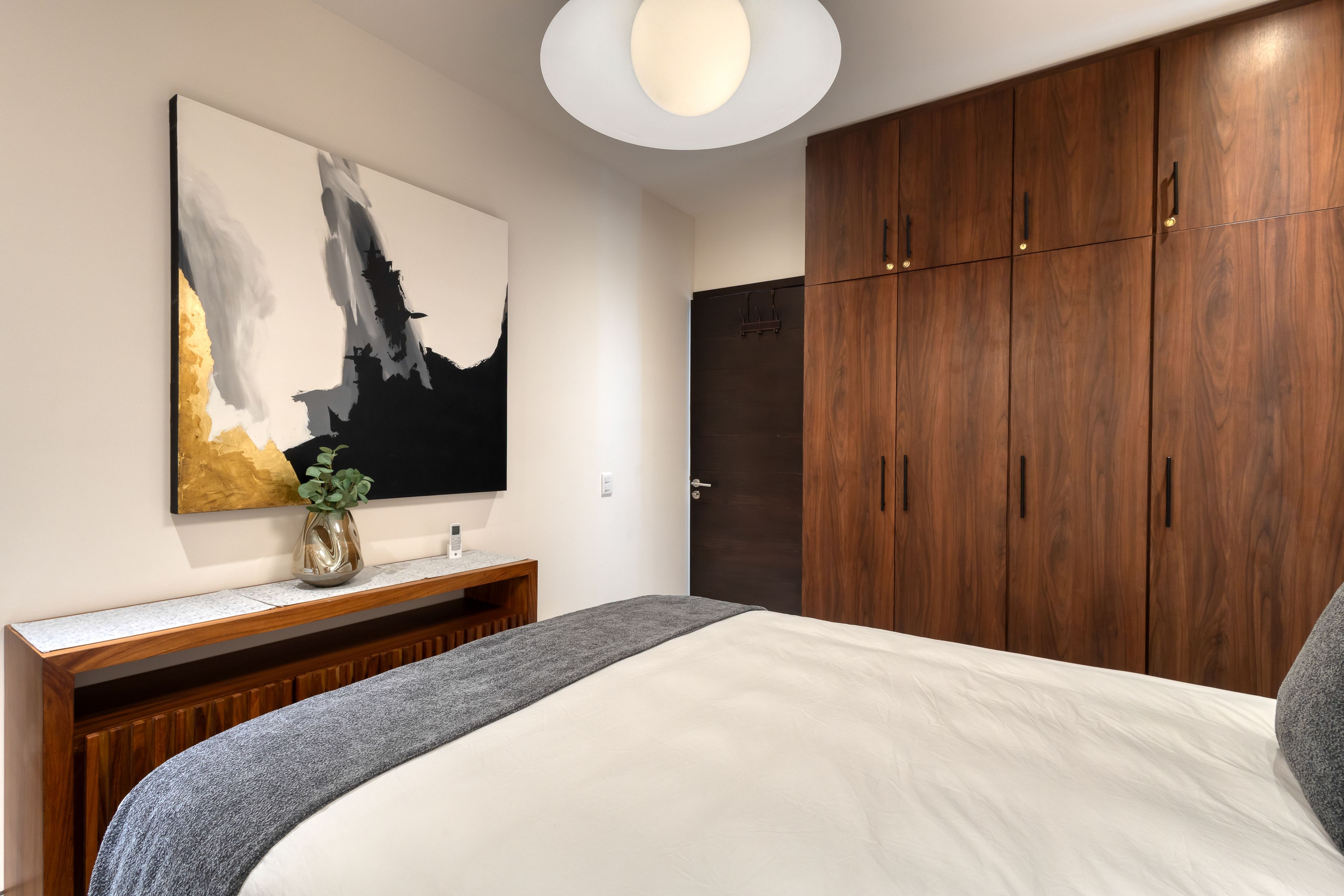 Une chambre à coucher avec un grand lit, une armoire en bois, un téléviseur fixé au mur et un tableau abstrait moderne.