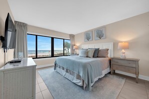 Ein Schlafzimmer mit einem großen Bett, einem Nachttisch, einem Fernseher und einem Fenster mit Blick auf den Strand.