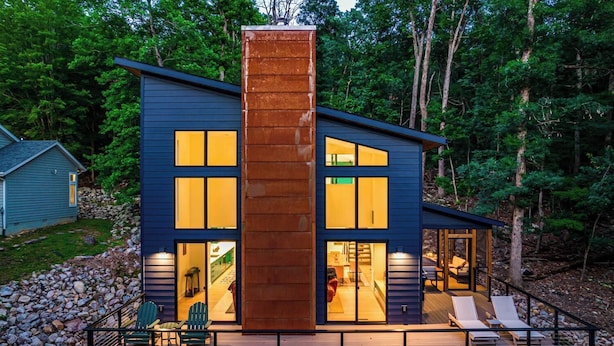A modern two-story house with a large central chimney, a deck with outdoor seating, and a stone pathway leading to the entrance.