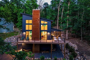 A modern two-story house with a prominent central chimney, large windows, and a deck with outdoor seating. - Luxury Log Cabin w Hot Tub & Sauna near Trail (McGaheysville)