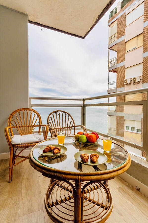 A balcony with wicker chairs, a glass table with croissants, fruit, and orange juice, overlooking a building and the sea. - Apartment on the beach - Paseo marítimo de Almería (Almería)