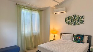 A bedroom with a bed, a bench, a nightstand with a lamp, an air conditioner, and a wall with tropical artwork. - Villa Saphir - Elegant family villa for 10 people with private pool in Occitanie, France (Saint-Jean-de-Cornies)