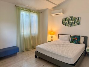 Ein Schlafzimmer mit Bett, Sitzbank, Nachttisch mit Lampe, Klimaanlage und Wand mit tropischer Kunst.