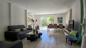 A modern living room with a sofa, coffee table, and chairs, a television, and a view through a glass door. - Villa Saphir - Elegant family villa for 10 people with private pool in Occitanie, France (Saint-Jean-de-Cornies)
