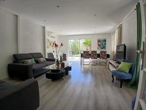 Ein modernes Wohnzimmer mit Sofa, Couchtisch und Sesseln, einem Fernseher und Blick durch eine Glastür.