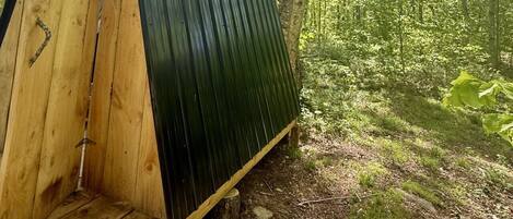 A wooden structure with a slanted roof, surrounded by a forested area.