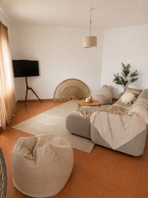 A living room with a sofa, a round bean bag, a TV on a tripod stand, a hanging light fixture, and a small table with candles.