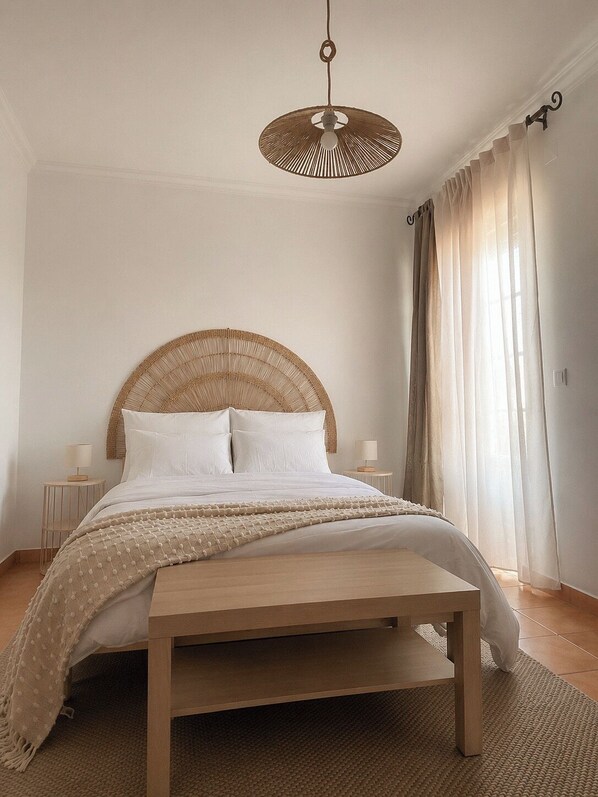 A bedroom with a wooden bed, a woven headboard, a hanging light fixture, and sheer curtains.